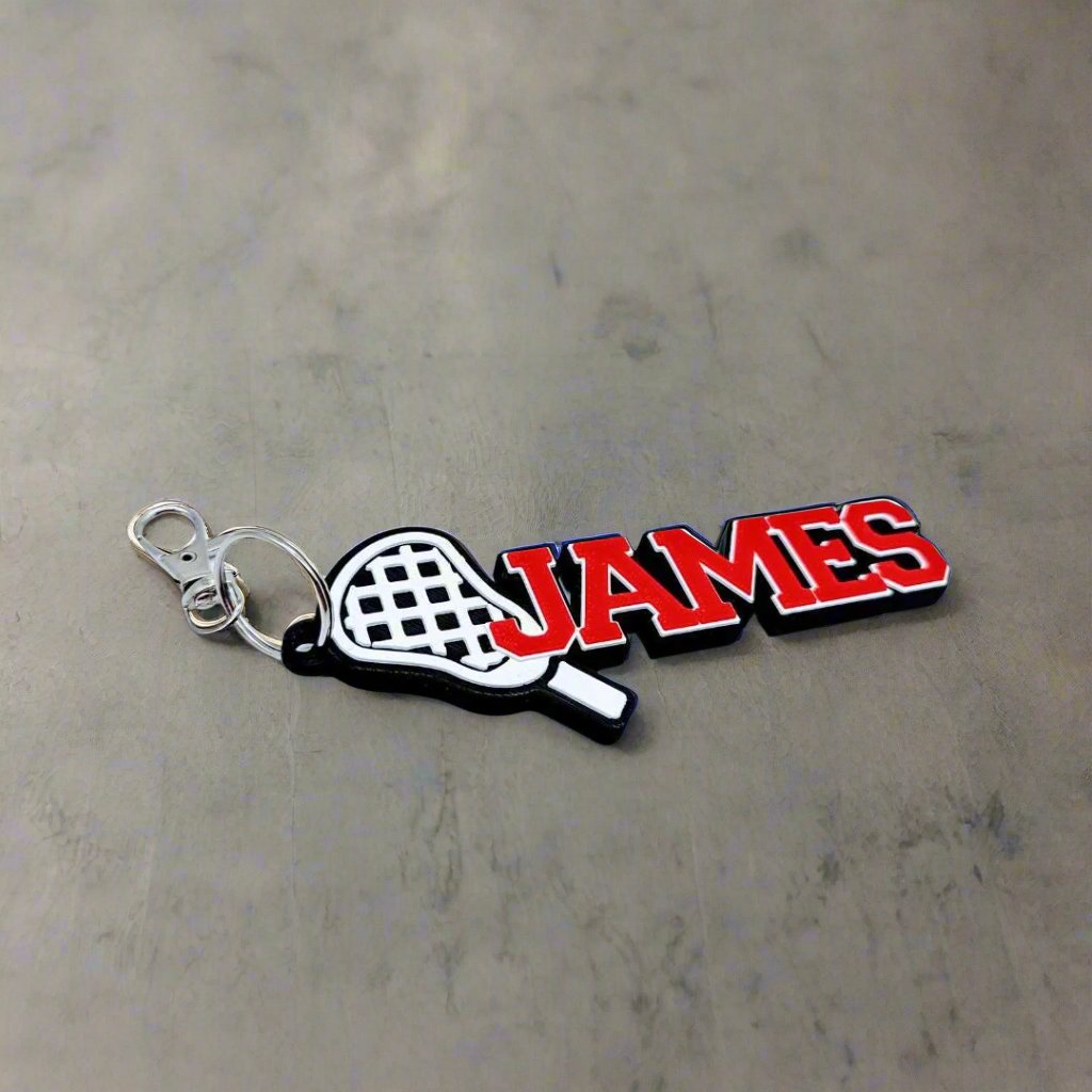 Red lacrosse name tag "JAMES" with metal clip on a concrete surface